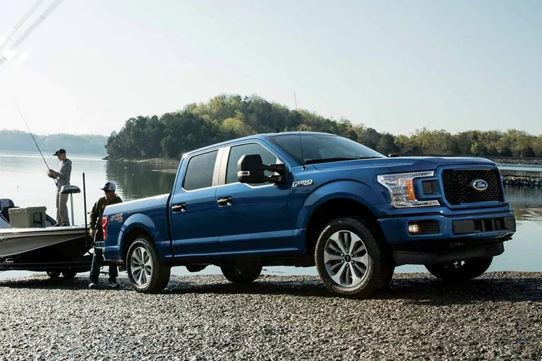 A blue Ford F-150 pickup truck parked by a lakeside, with a person standing near the tailgate and preparing a boat for launch. The serene lake and distant tree-lined horizon can be seen in the background on a sunny day.