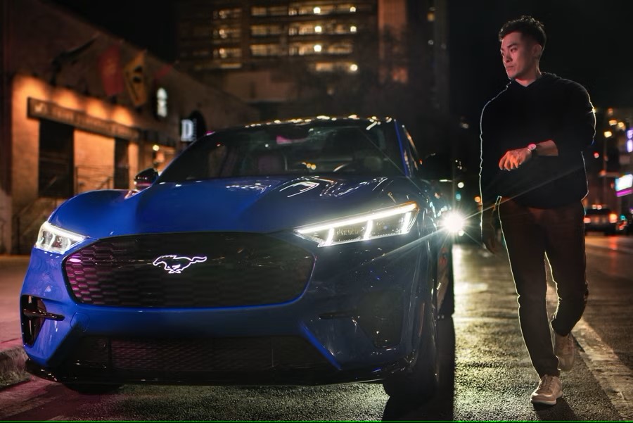 A man in dark clothing stands on a city street at night, illuminated by the headlights of a sleek blue Ford Mustang Match-E electric car. The city lights and buildings form a dim backdrop, adding ambiance to the scene.